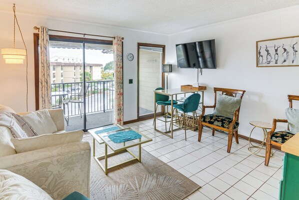 A living room with a sofa, a coffee table, a TV, and a balcony with a view of buildings. - Pool, Spa & Dining: Beach Condo in Hilton Head! (Hilton Head)