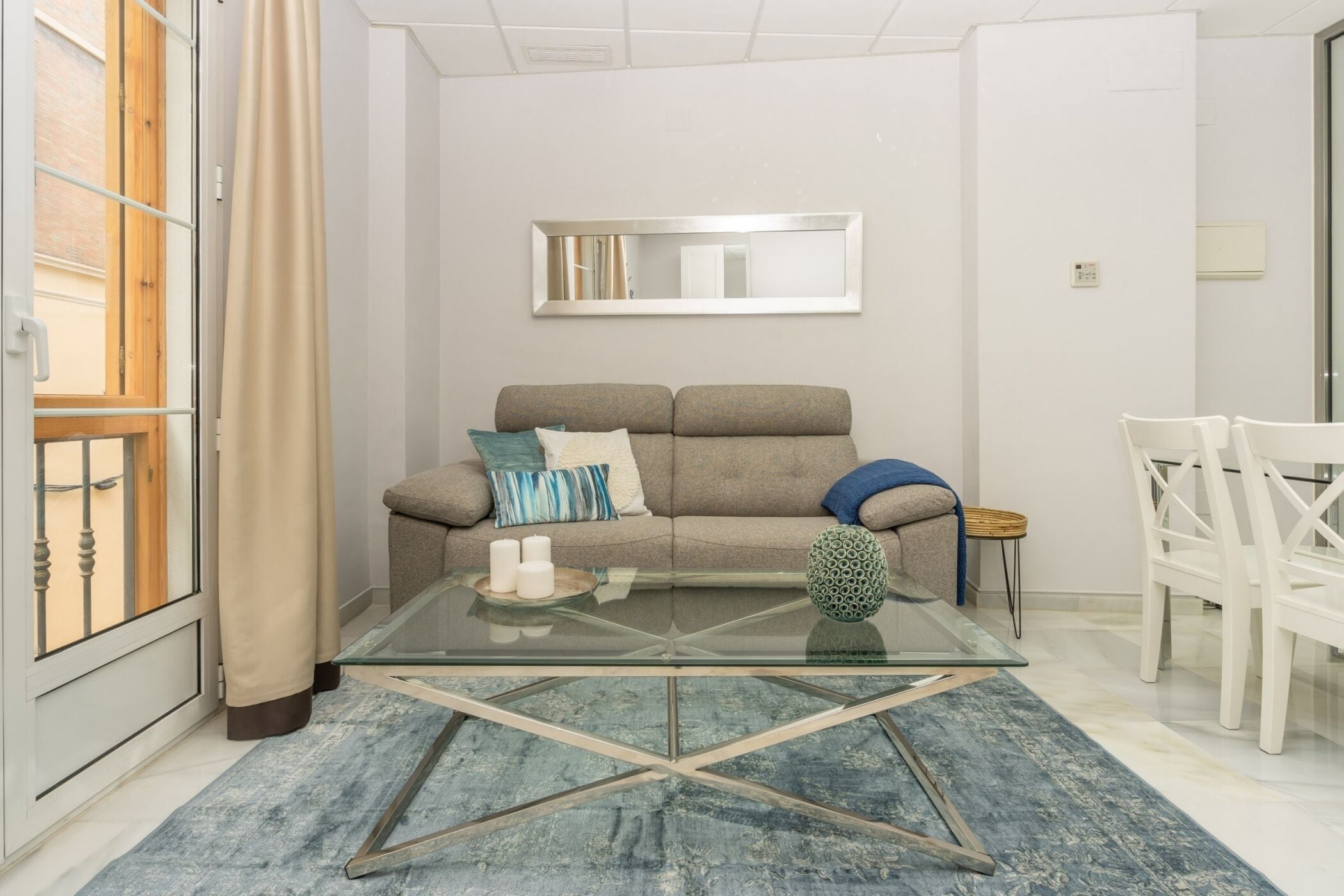 A modern living room with a glass coffee table, a sofa with cushions, a small side table, and a white chair.