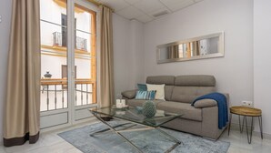 A modern living room with a glass coffee table, a grey sofa with cushions, a blue rug, and a balcony with a view of buildings. - iloftmalaga Strachan-Larios (Málaga)
