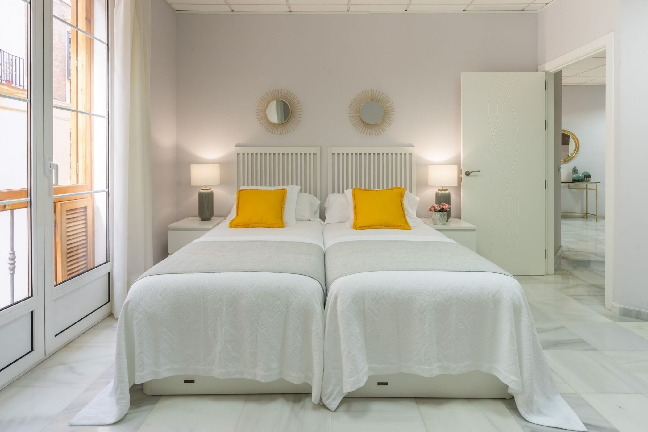 Un lit double avec une literie blanche et deux oreillers jaunes, de part et d’autre de deux tables de chevet surmontées de lampes, dans une pièce dotée d’une grande fenêtre et d’une porte donnant sur une autre pièce.