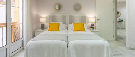 Un lit double avec une literie blanche et deux oreillers jaunes, de part et d’autre de deux tables de chevet surmontées de lampes, dans une pièce dotée d’une grande fenêtre et d’une porte donnant sur une autre pièce.