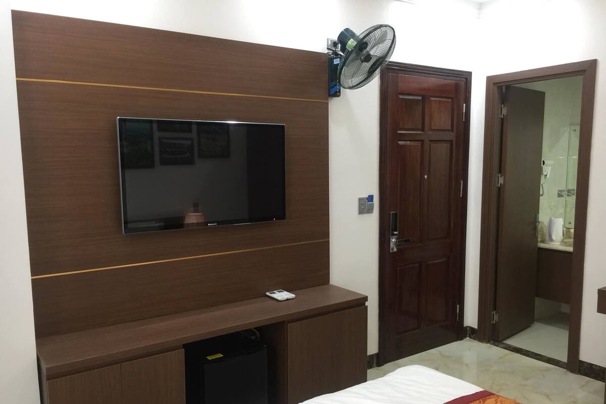 Quarto de hotel com televisão de tela plana fixada em painel de madeira, pequena geladeira, mesa de cabeceira e cama com manta estampada.