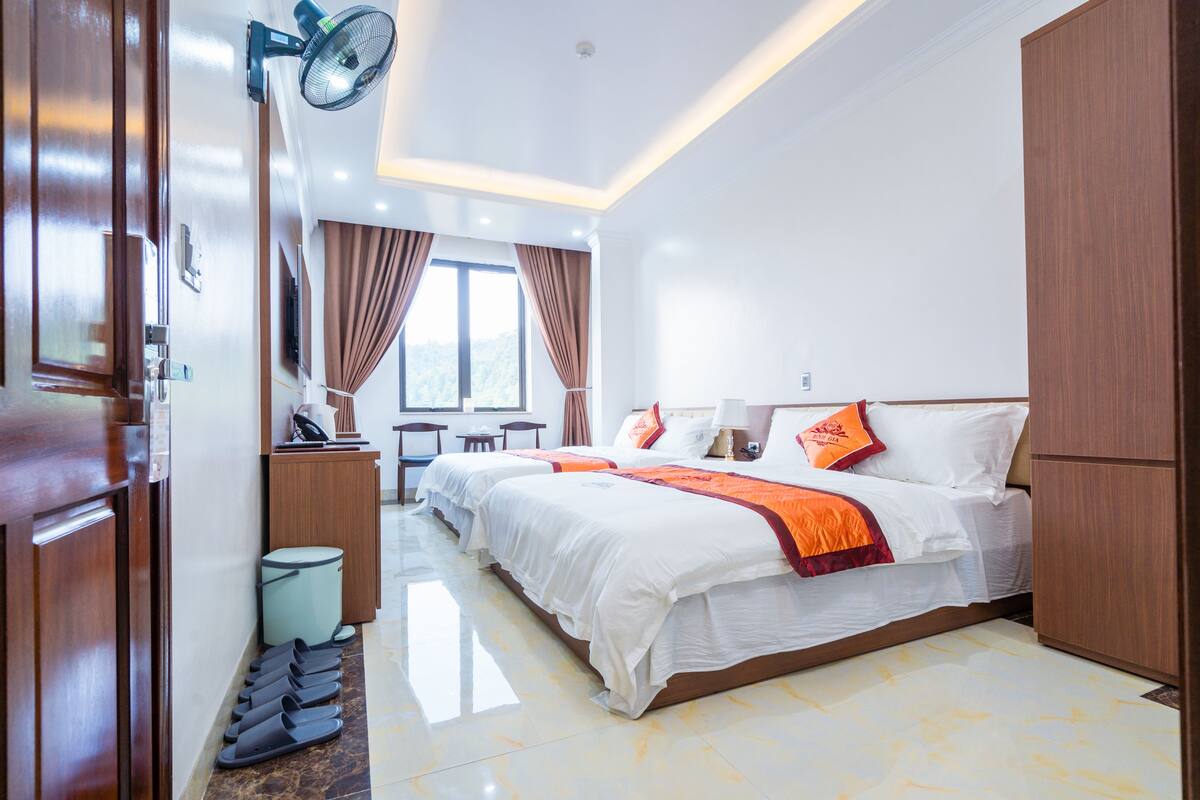 Quarto de hotel com uma cama grande, uma mesa e uma cadeira. Há uma janela com cortinas, um ventilador e uma lixeira.