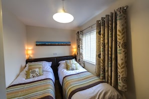 Quarto de hotel com duas camas de solteiro, mesa de cabeceira com abajur, um quadro na parede e janela com cortinas.