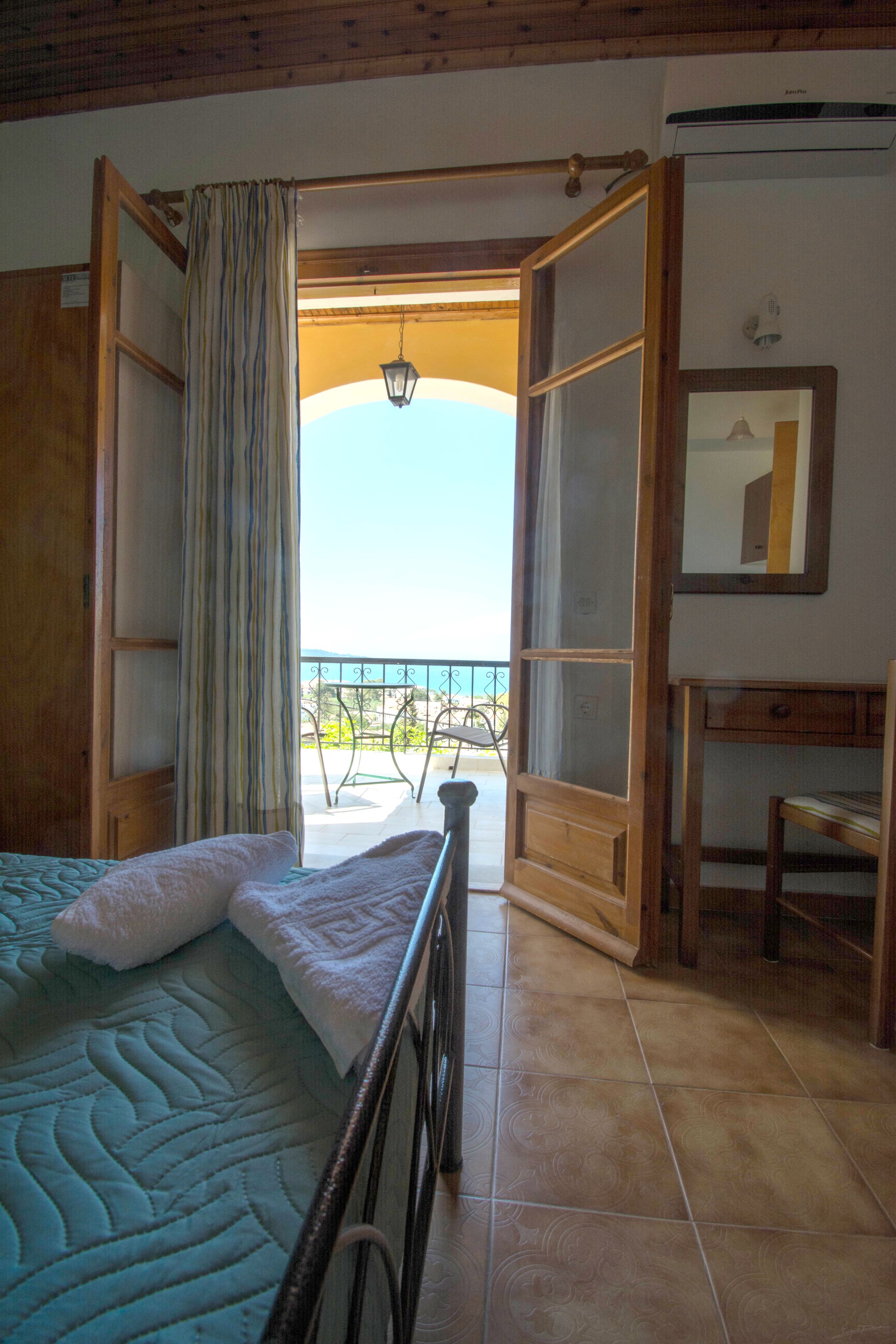 A room with a bed, a wooden desk, and an open door leading to a balcony.
