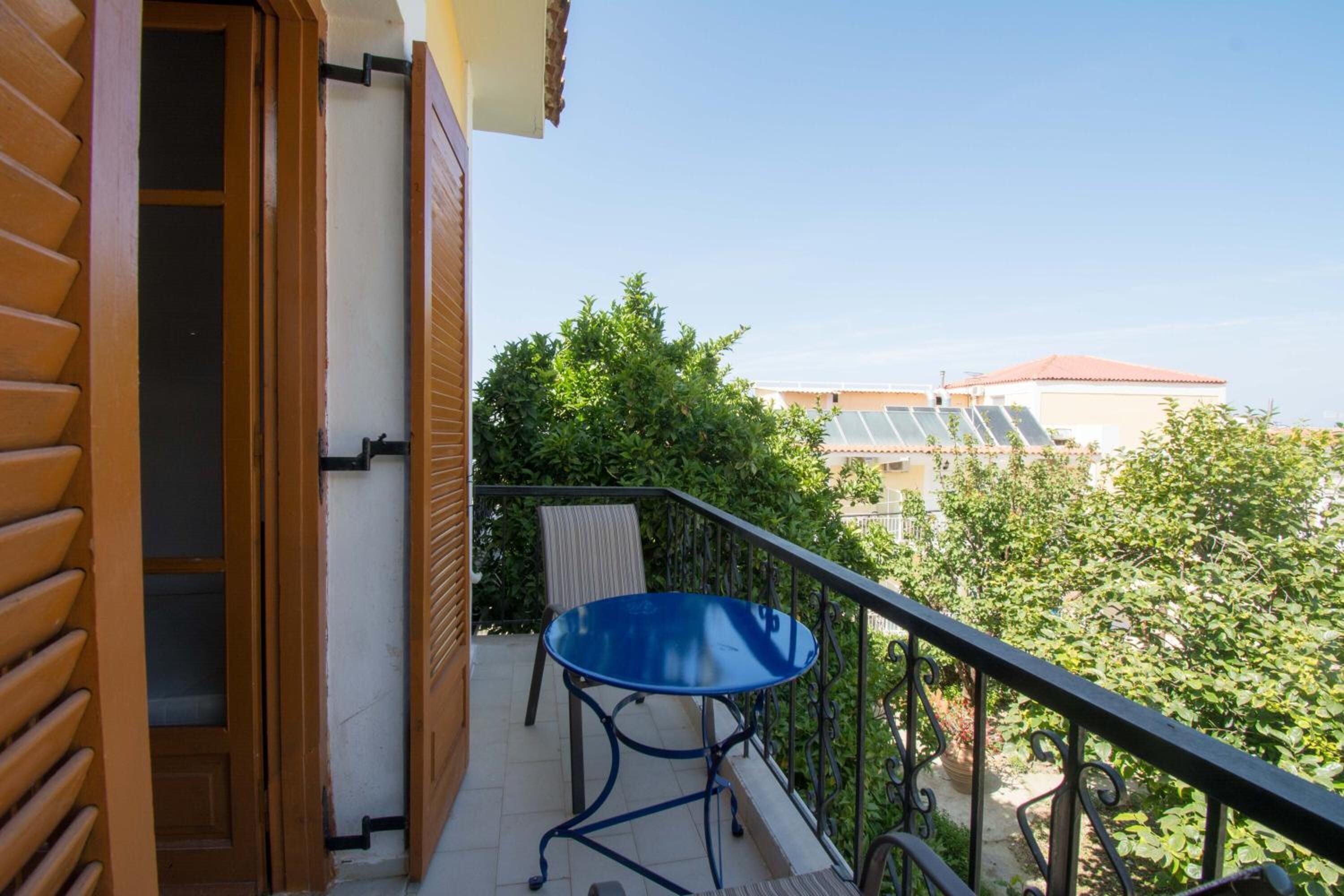 A balcony with a blue table and chair, a wooden door, and a view of greenery and buildings in the background.