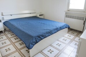A single bed with a blue quilt, a white headboard, and a white nightstand. The floor has patterned tiles. - M224 - Marcelli, three-room apartment with garden 150m from the sea (Marcelli)
