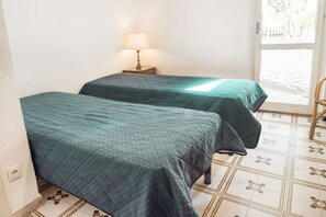A room with two beds, a nightstand with a lamp, and a window with a view of the outside.
