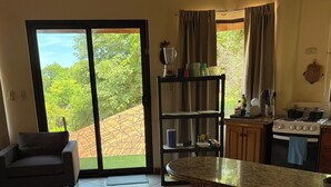 A cozy living room with a sofa, a wooden bench, and a dining table. There is a kitchen area with a stove, shelves, and a view of greenery outside. - Your Comfortable, Safe, Updated Villa Retreat for 2 to 6 Guests in Playa Hermosa (Playa Hermosa)