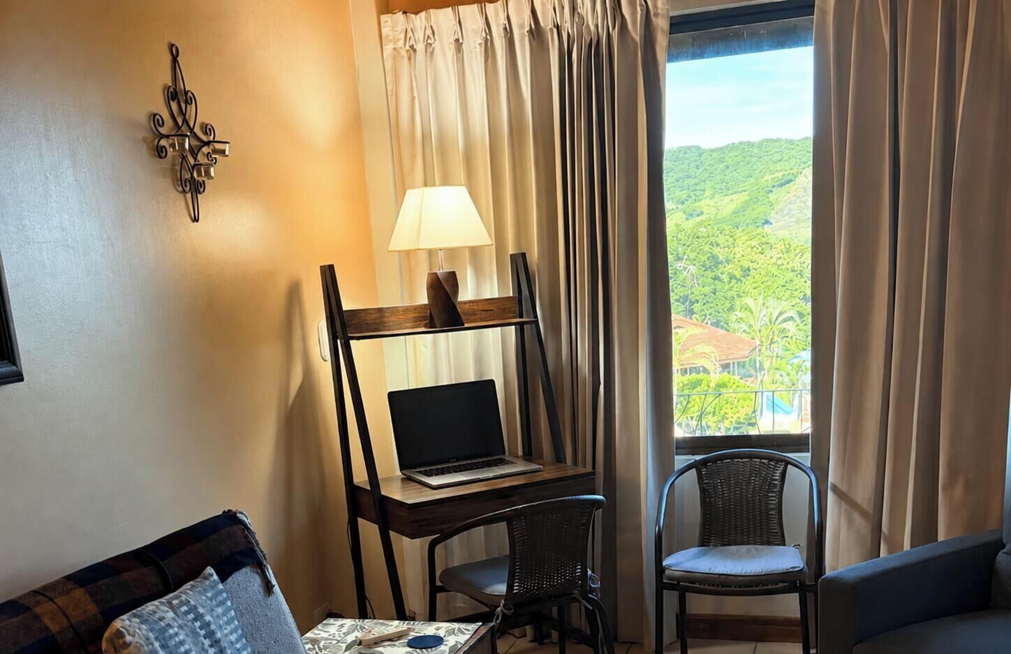 Una stanza con scrivania, computer portatile, lampada e vista su un paesaggio verde attraverso la finestra.