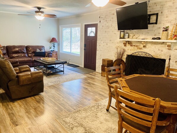 A living room with a brown leather sofa, a wooden dining table with chairs, a ceiling fan, and a fireplace with a TV mounted above it.