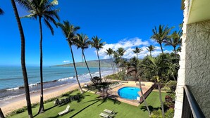 Zona de piscina con vista a la playa y al océano, rodeada de palmeras y un edificio blanco.