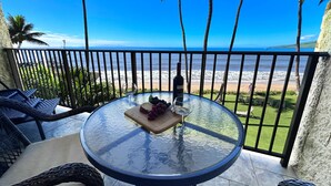 Una mesa redonda de vidrio con una botella de vino, una copa y una tabla de madera con uvas en un balcón con vistas a la playa.