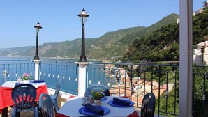 A terrace with a table set for dining, overlooking a coastal area with hills and buildings. - Sea Side Rooms (Scilla)