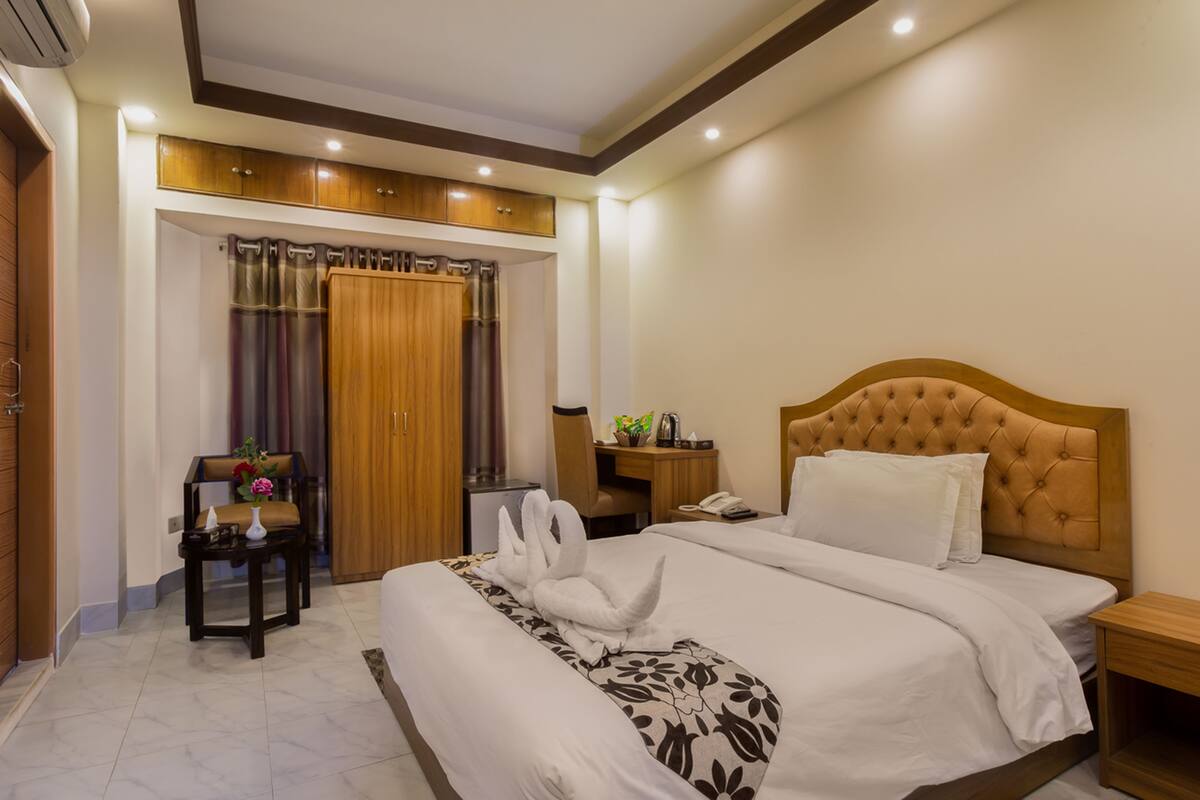 A hotel room with a large bed, a wooden headboard, a desk with a chair, a wardrobe, and a bathroom with a shower curtain.
