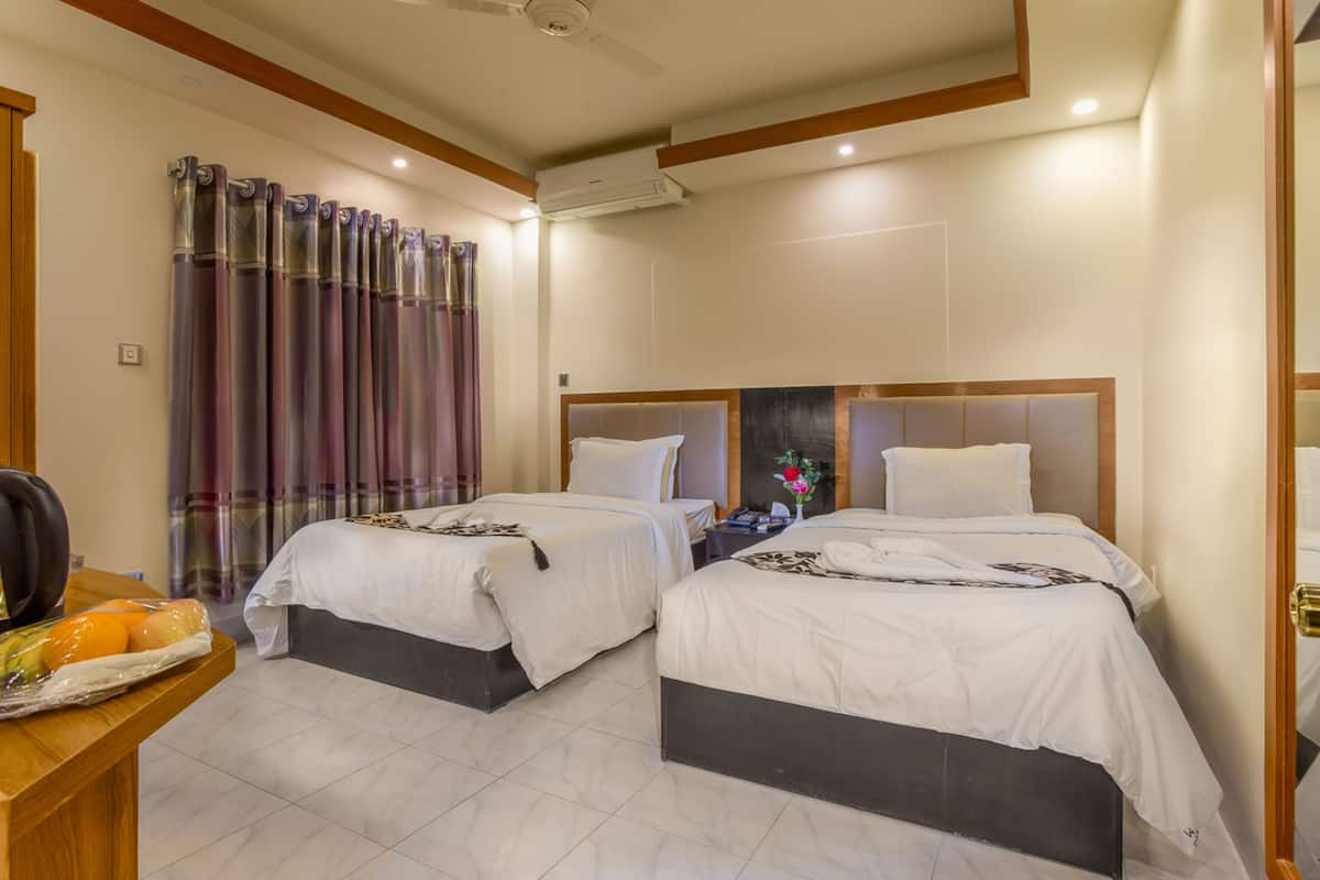 A hotel room with two beds, a wooden desk with a fruit basket, and a window with purple curtains.
