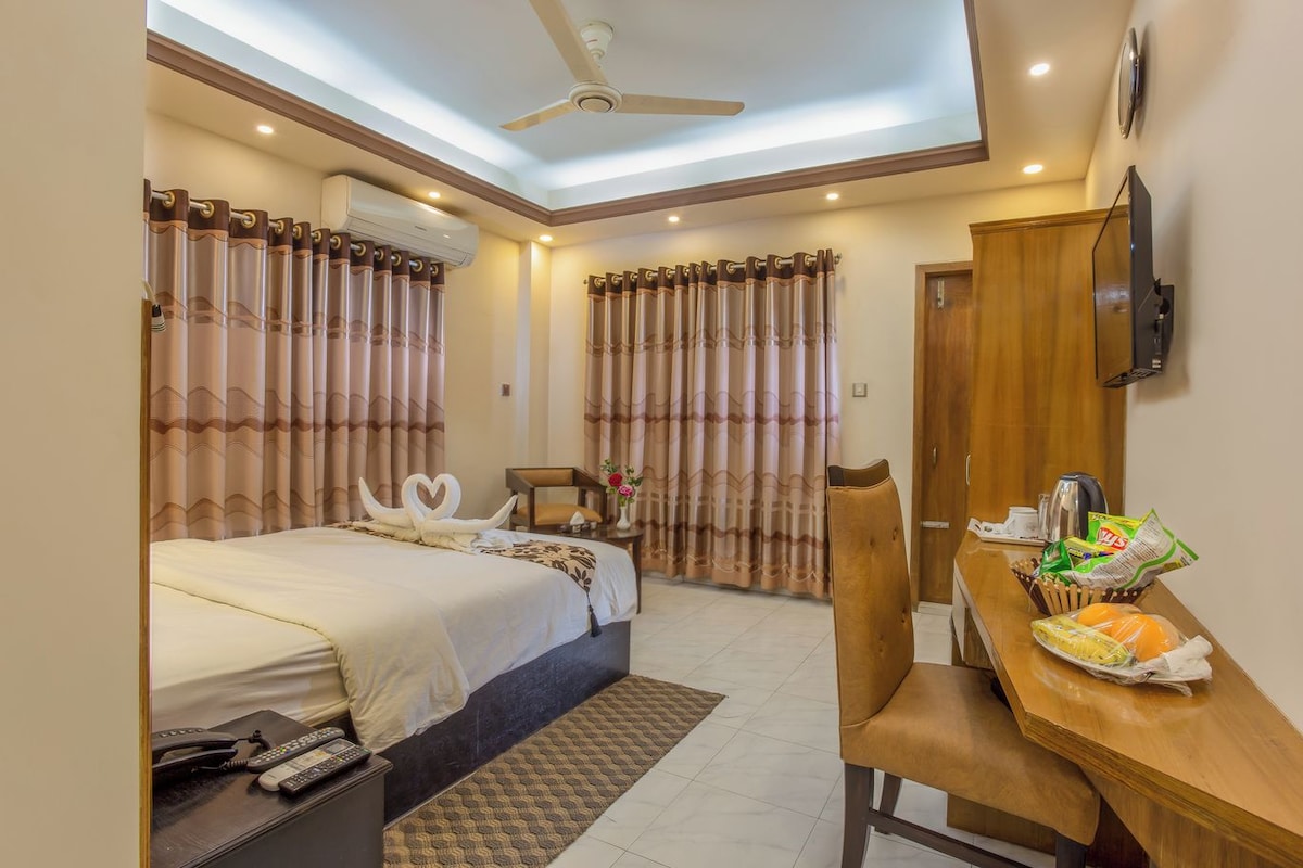 A hotel room with a bed, a desk with breakfast items, a chair, a TV, and an air conditioning unit.