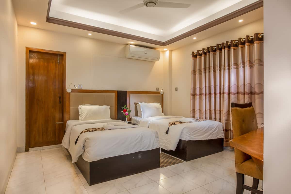 A hotel room with two beds, a wooden door, an air conditioning unit, a ceiling fan, and a dining table with a chair.