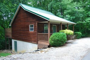 Situated in a wooded area, this cabin features a gabled roof, front porch, and a small entrance.