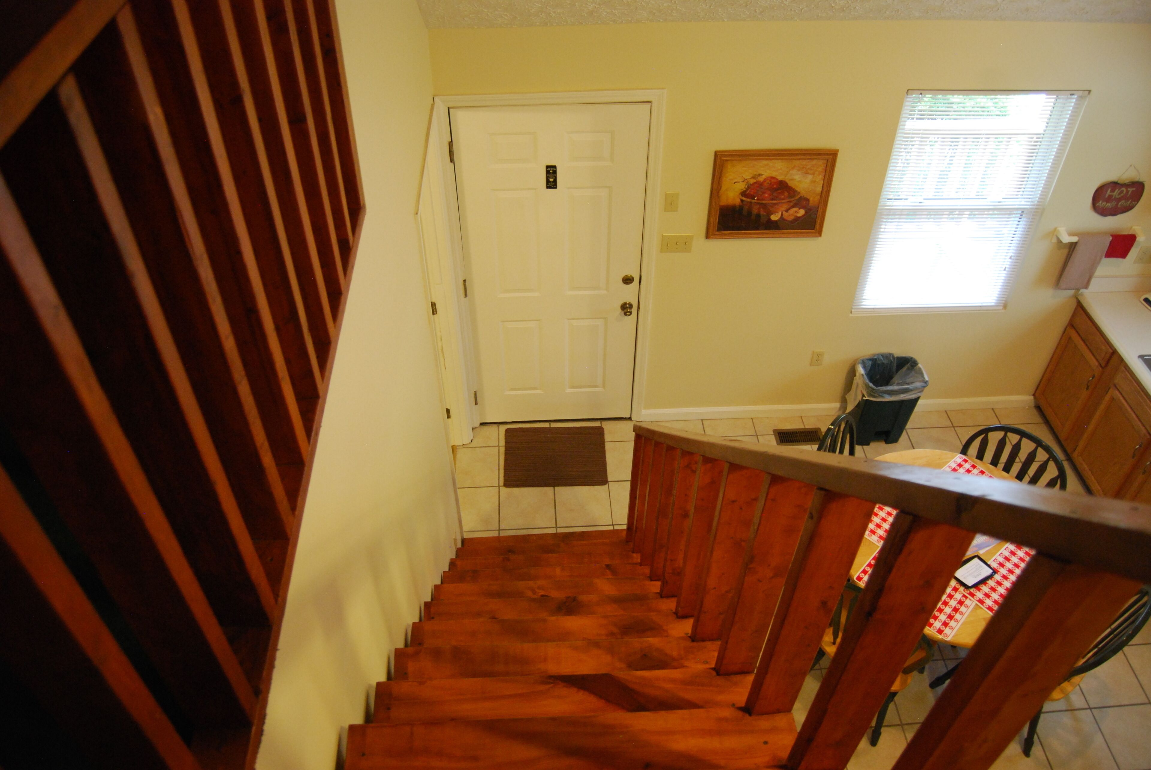 A wooden staircase leading to a white door, a window with blinds, and a chair with a checkered tablecloth.