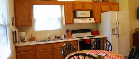 Una cocina con armarios de madera, microondas, estufa y refrigerador. Hay una mesa de comedor con sillas y mantel a cuadros rojo y blanco.