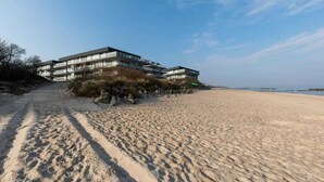 Een gebouw aan het strand met meerdere balkons, een zandstrand voor op de foto en een heldere lucht.