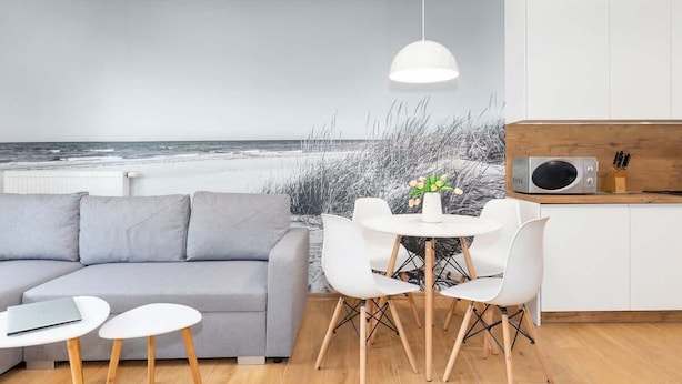 Een moderne woonkamer met een grijze bank, witte stoelen, een klein rond tafeltje en een hanglamp. Een keuken met een magnetron en houten kastjes is zichtbaar.
