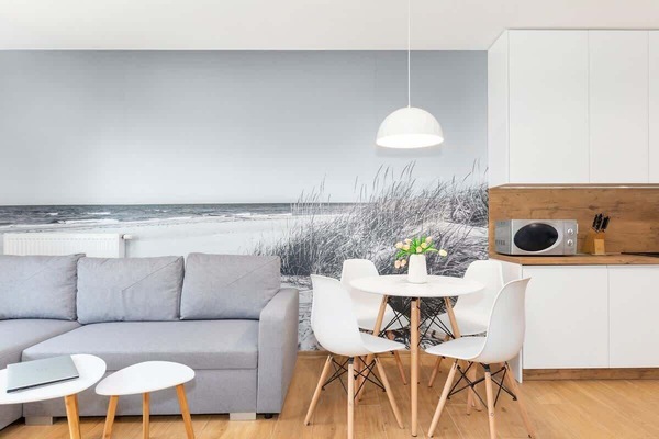 Bright living room with a grey sofa, round dining table, and an open kitchenette with white cabinets and wood accents.