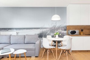 A modern living room with a gray sofa, white chairs, a small round table, and a pendant light. A kitchen area with a microwave and wooden cabinets is visible. - Bright Apartment with Sea View | Close to the Beach and Parking (Dziwnów)