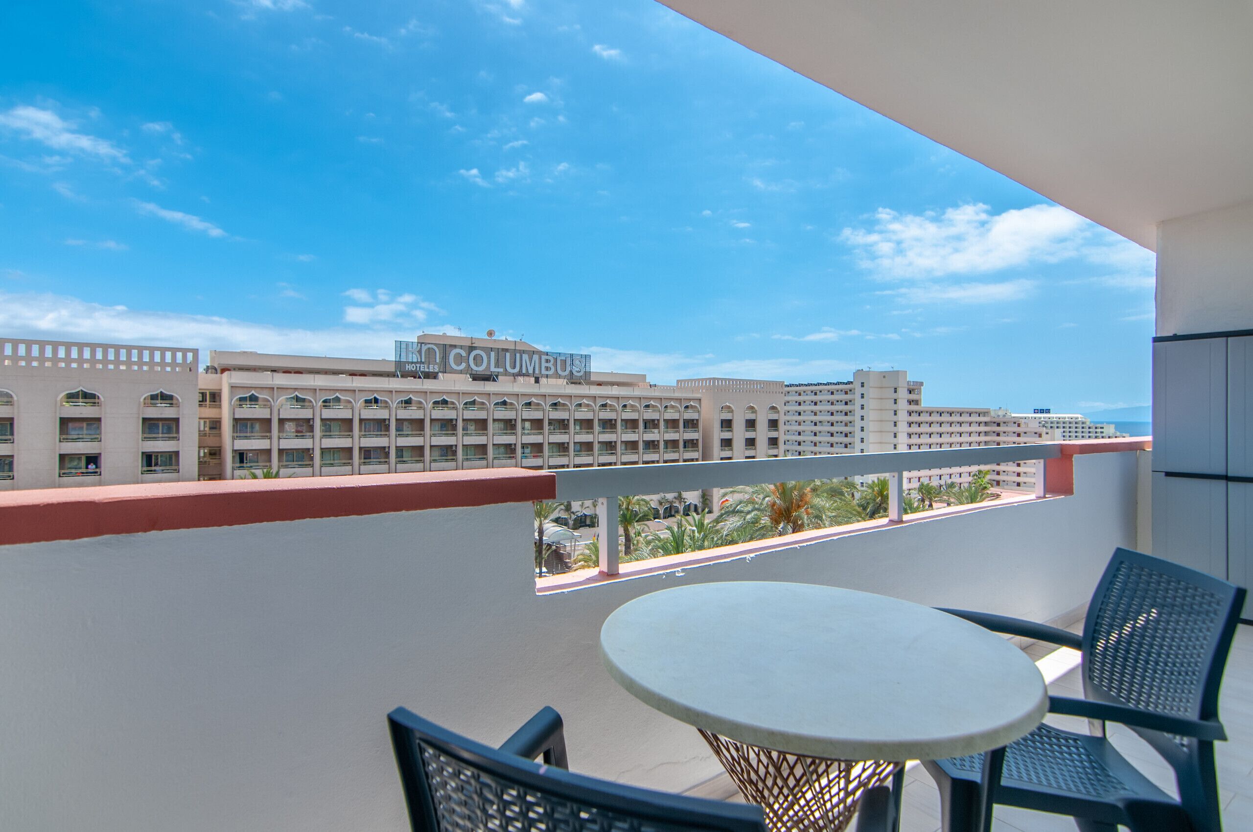 A balcony with a round table and chairs, overlooking a hotel with a sign reading "HOTEL COLUMBUS".