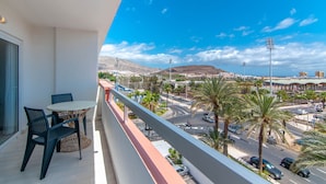 A balcony with a table and chairs, overlooking a busy street with palm trees and a mountain in the background. - Los Tajinastes 1A034 (Playa de Las Americas)