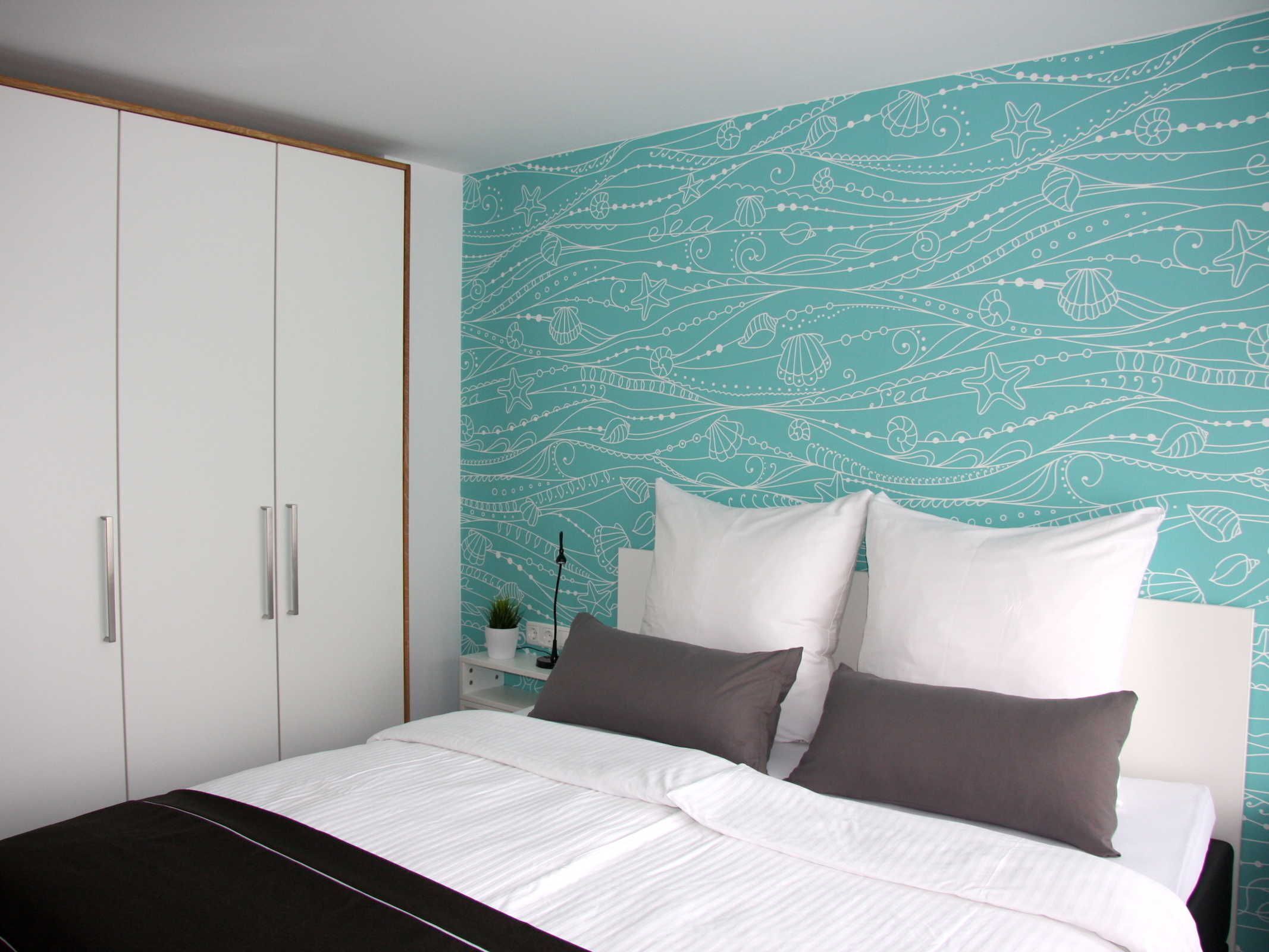 A bedroom with a bed, white and dark grey pillows, and a teal patterned wall.