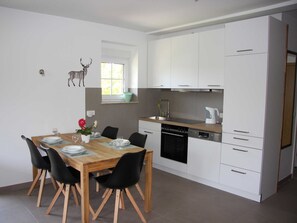 A modern kitchen with white cabinets, a wooden dining table, black chairs, and a small window with a view of greenery.