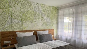 Un dormitorio con cabecera de madera, cama con ropa de cama blanca y gris, pared con estampado de hojas verdes y una ventana con cortinas traslúcidas.