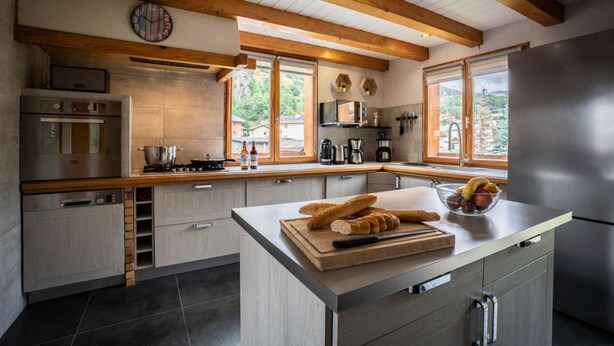 A modern kitchen with wooden beams, stainless steel appliances, and a large island with bread and fruit.
