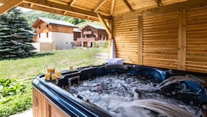 A wooden cabin with a hot tub, outdoor seating, and a view of a mountain and buildings.