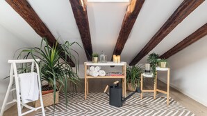A cozy attic room with exposed wooden beams, a skylight, a white ladder, and a wooden shelf with plants and books.