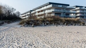 Un edificio moderno de varios pisos con balcones, ubicado en una playa de arena con vegetación escasa.