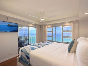 Une chambre dotée d’un grand lit, d’un ventilateur de plafond et offrant une vue sur l’océan grâce à de grandes fenêtres.
