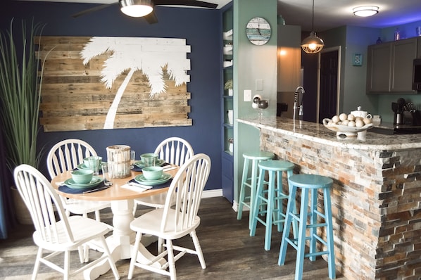 A dining area with a wooden table and white chairs, a kitchen with teal accents, and a decorative wall piece.