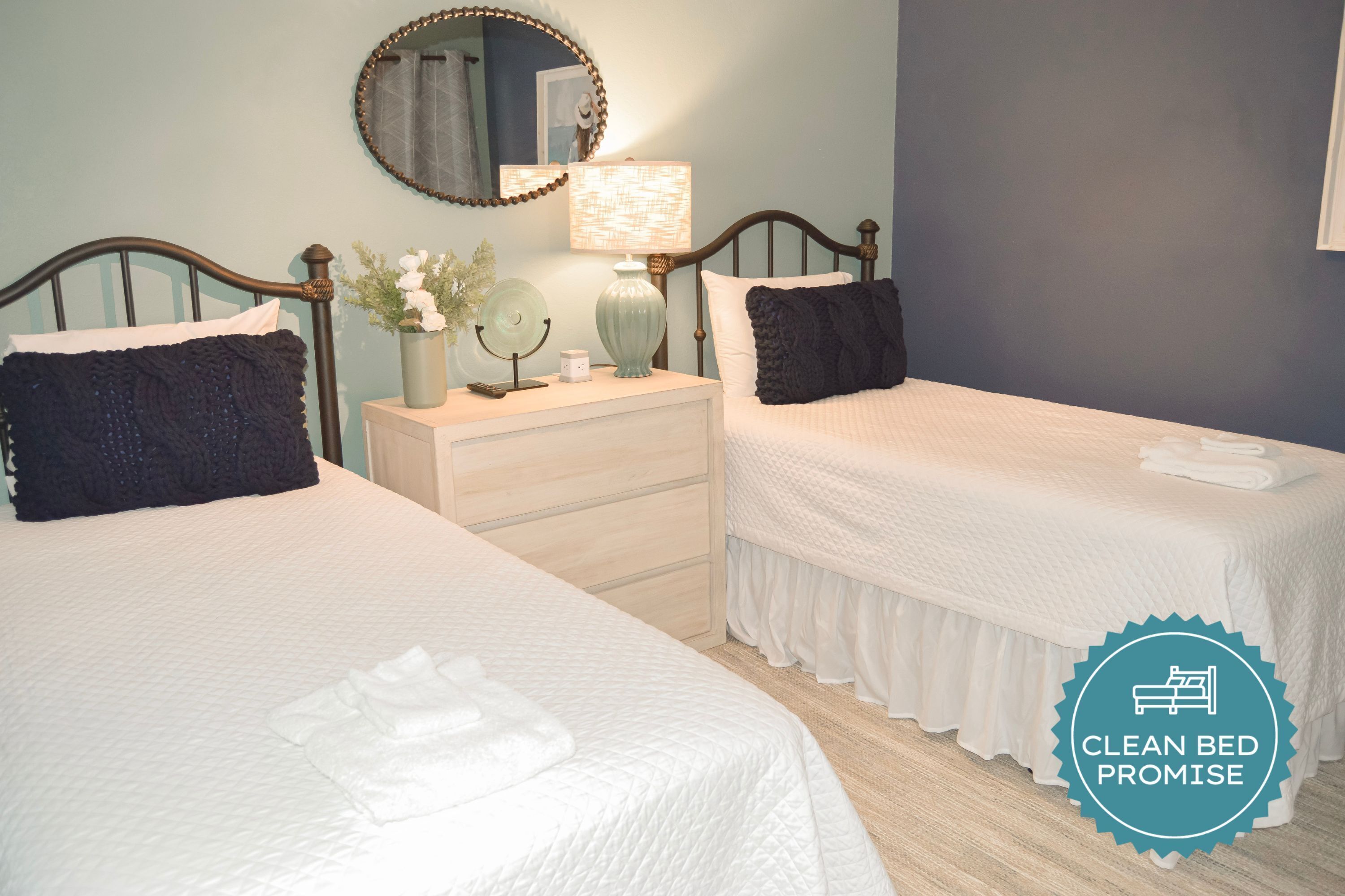 A clean, well-lit bedroom with two beds, a bedside table with a lamp, a mirror, and a decorative vase.