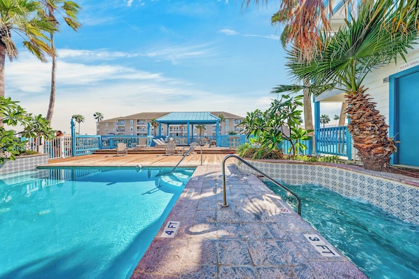 A swimming pool with a blue and white tiled edge, a wooden deck, and a building with a blue roof in the background.