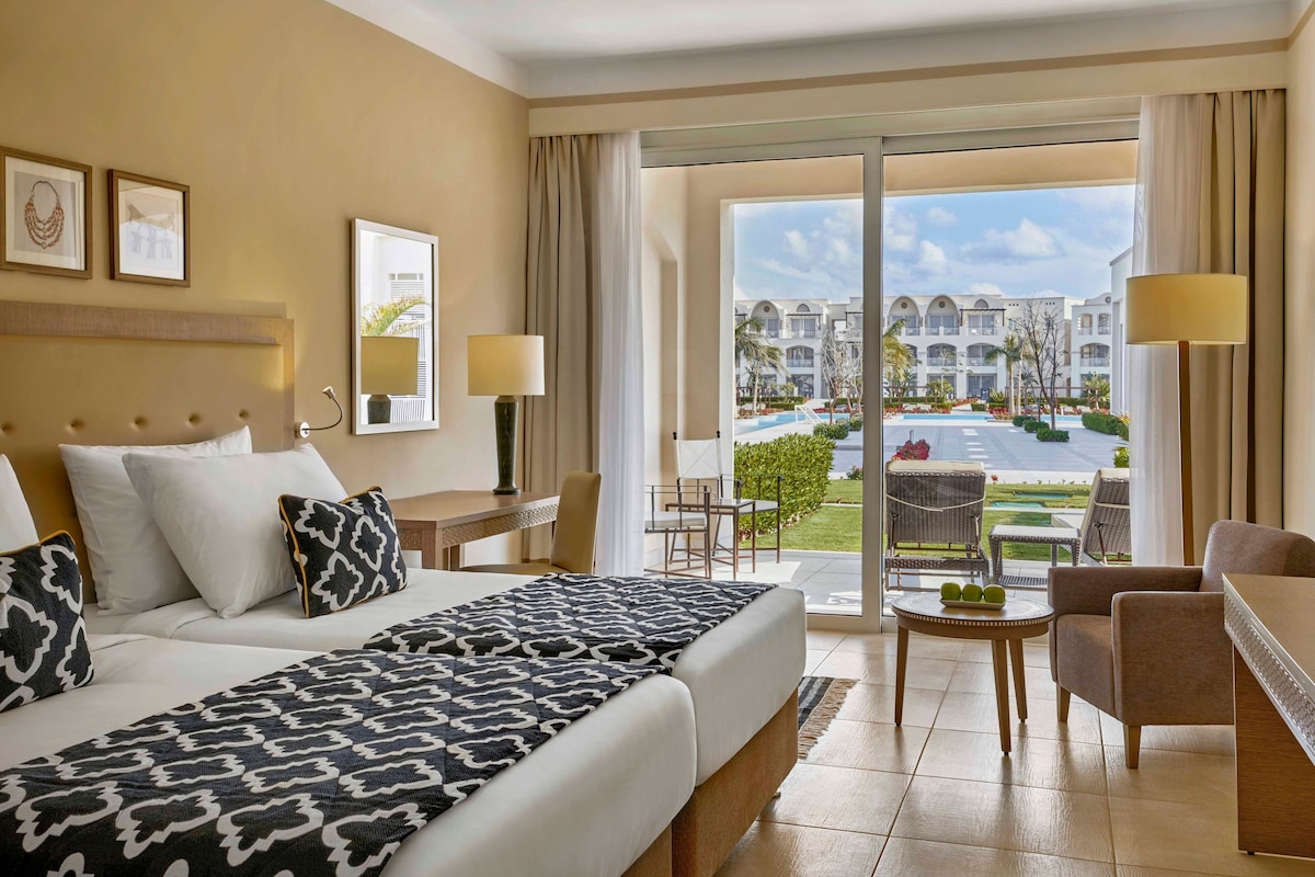 Une chambre d’hôtel avec un lit, un bureau, une chaise, une table avec des fruits, et une vue sur l’hôtel et la piscine.