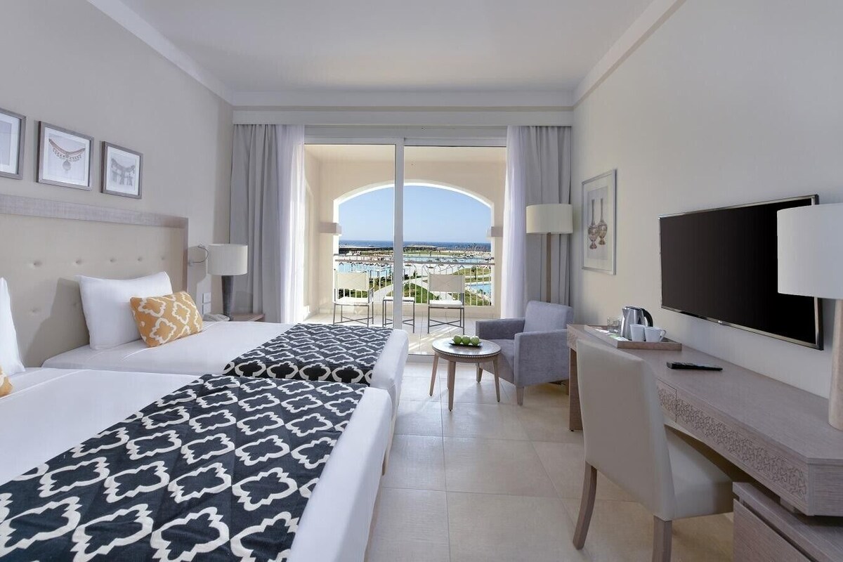 Une chambre d’hôtel avec un grand lit, un bureau avec une télévision, un balcon avec vue et un coin repas.