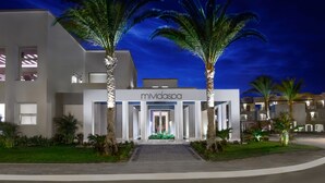 Une entrée d’hôtel moderne, avec une enseigne indiquant « mividaspa », des palmiers et un chemin bien éclairé menant à l’entrée.