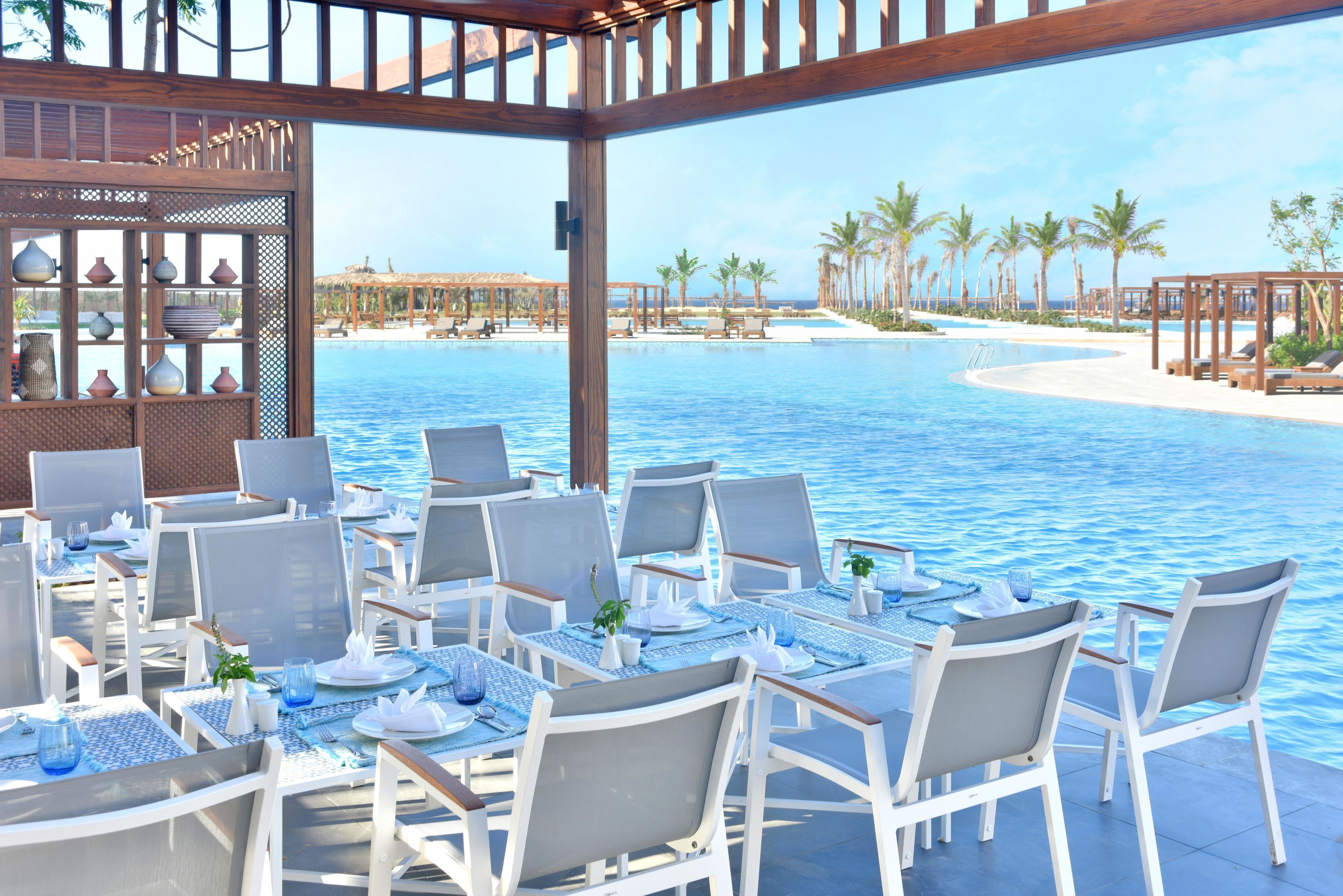 Un espace de restauration aménagé au bord de la piscine, avec des tables et des chaises, offrant une vue sur une piscine bordée de palmiers et une plage en arrière-plan.