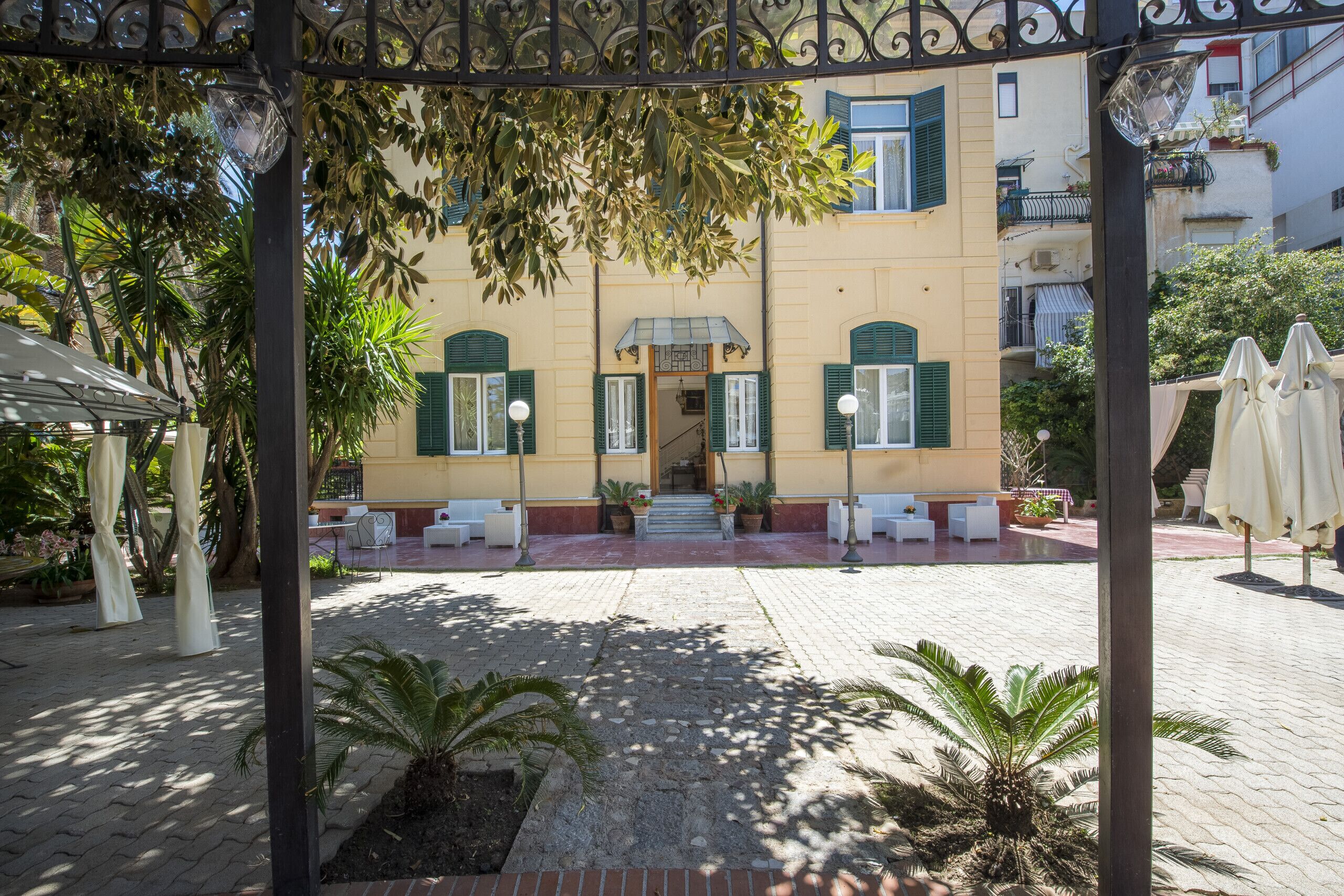 Un cortile con un edificio caratterizzato da un ingresso centrale, piante in vaso, posti a sedere all'aperto e ombrelloni.