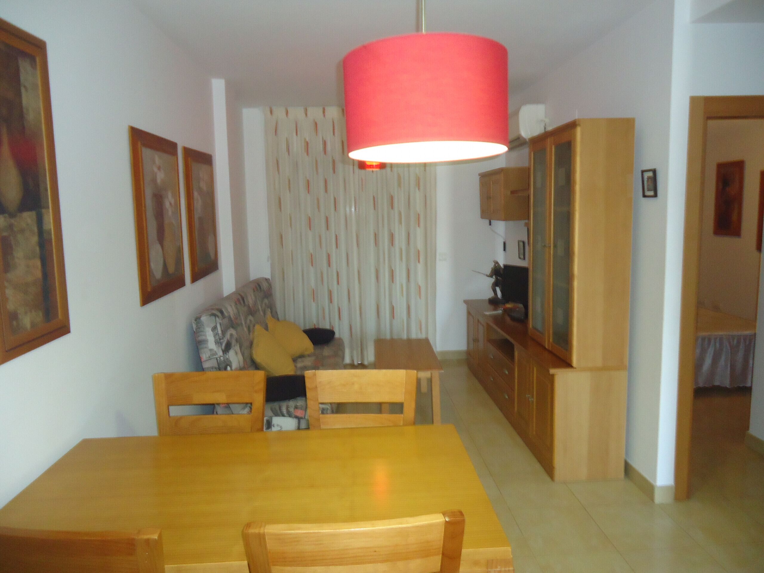 A dining area with a wooden table and chairs, a red hanging lamp, a sofa with a yellow cushion, and a patterned curtain.