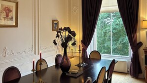 Ein Esszimmer mit einem langen Tisch, Stühlen, einer Chandelier, einer Vase mit Blumen und einem Fenster mit Vorhängen.
