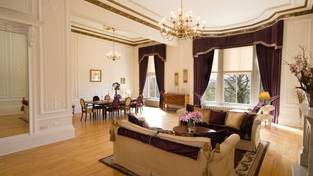 Ein geräumiges Wohnzimmer mit Essbereich, einer Chandelüster und großen, mit Vorhängen versehenen Fenstern.